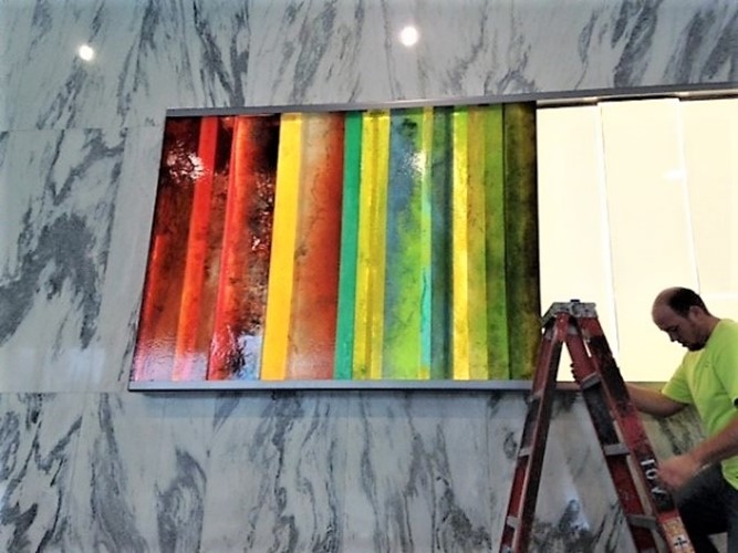 person, likely an artist or installer, working on hanging a large, colorful abstract artwork with vertical stripes on a wall