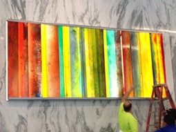 person, likely an artist or installer, working on hanging a large, colorful abstract artwork with vertical stripes on a wall